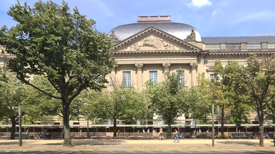 Foto av fasaden på Staatsbibliothek zu Berlin. Träd och grönska i förgrunden.