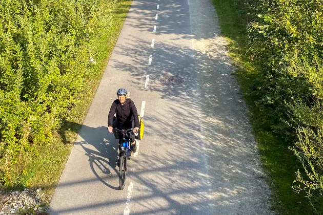 Foto taget uppifrån. Bilden visar en lång cykelväg omgiven av skog och en kvinna som cyklar längs den.