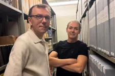 Två män som står mellan en massa bokhyllor på ett arkiv.