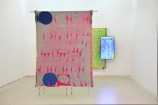 En färgstark väv i rosa, grått och blått hänger från taket i bakgrunden ytterligare en väv, grön, tillsammans med en digital skärm som visar en video.