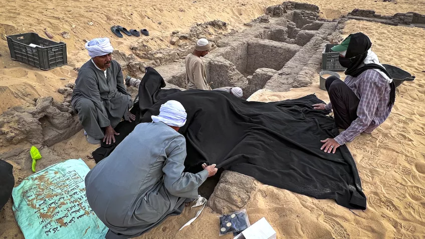 Foto från en utgrävning i den egyptiska öknen, där tre personer håller ett svart skynke ovanför ett utgrävt magasin.